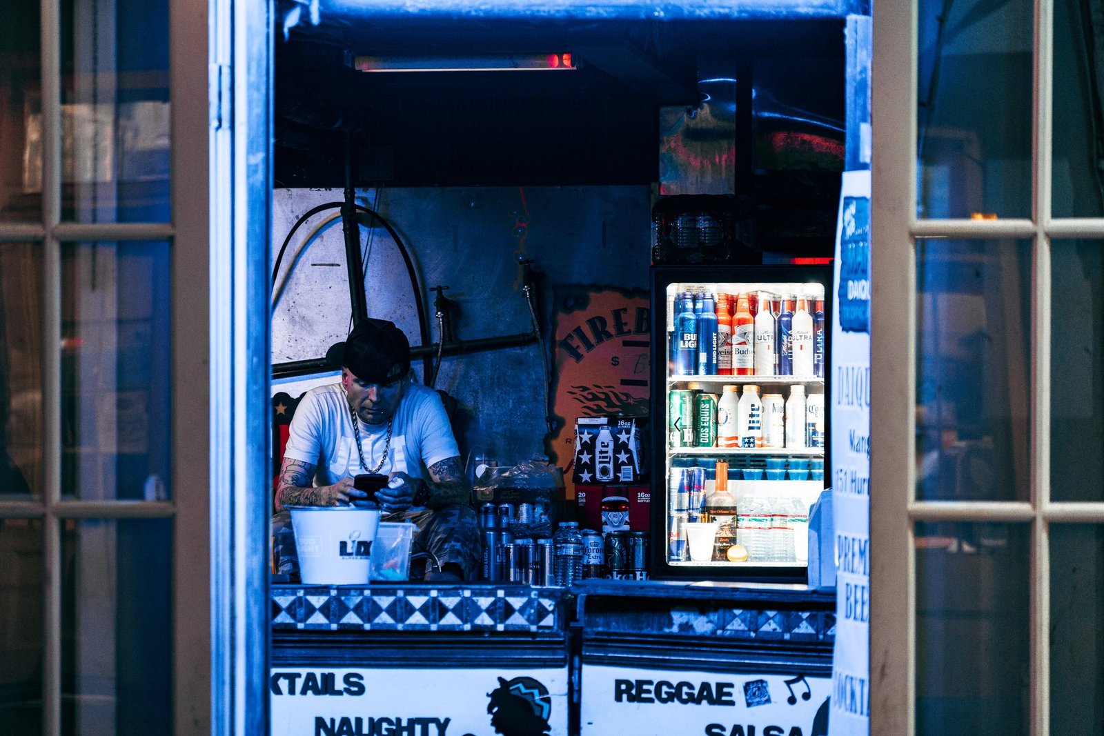Man on a phone inside a blue-lit daiquiri shop window, beer cooler glowing behind him, New Orleans, Louisiana